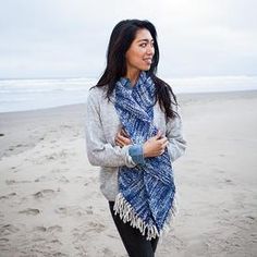 a woman standing on top of a sandy beach next to the ocean wearing a blue and white scarf