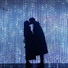 two people standing in front of a wall with lights on it and one person kissing the other