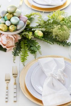 an easter table setting with flowers, eggs and greenery on the plates in gold rimmed dishes