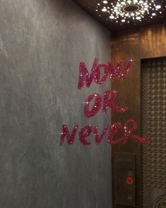 the words now or never are spelled in bright pink glitters on a gray wall