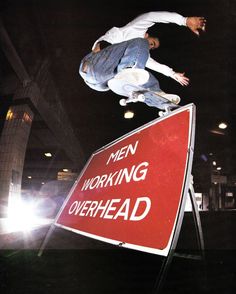 a man on a skateboard jumping over a sign that says men working overhead in front of him