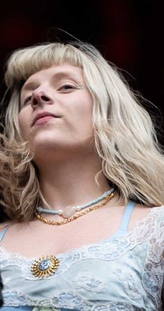 a woman with blonde hair wearing a blue dress and gold necklace looking up into the sky