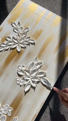 someone is painting flowers on a canvas with gold and white paint while pointing at it