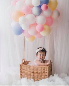 a baby sitting in a basket with balloons floating above her and on the floor next to it