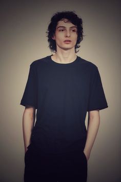 a young man with curly hair wearing a black t - shirt and standing in front of a gray wall