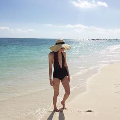 a woman standing on the beach wearing a hat and black swimsuit with her hands in her pockets