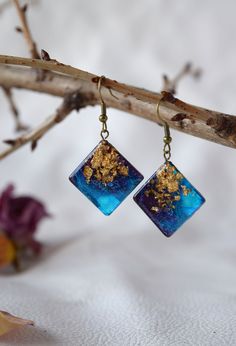 two square blue and gold earrings hanging from a twig with flowers in the background