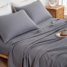 a bed with grey sheets and pillows on it next to a basket full of coffee