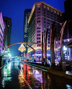 a city street at night with lights reflecting off the wet pavement and tall buildings in the background