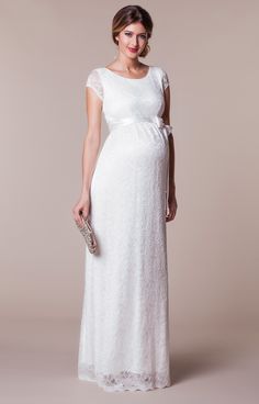 a pregnant woman wearing a white gown and holding a clutch in one hand, standing against a beige background