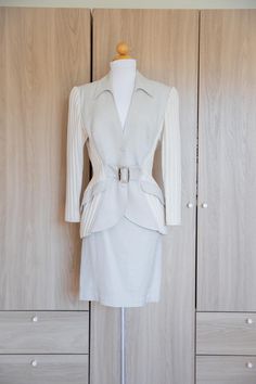 D E T A I L S * 90s Thierry Mugler Mix Fabric Skirt Suit Set * jacket features gorgeous contrasting knit sleeves and mid-section in an off-white shade paired with a light gray linen fabric * pencil high waist skirt with zipper closure, snap button and hook and eye * jacket has a curved down pointed collar  * jacket is nipped at the waist with faux pockets that curve down and an attached silver buckle belt to cinch the waist which gives way to a sculptural peplum with high-low cascading hem * jacket has light shoulder padding  *  jacket has front and back waist darting * material isa mix of linen, cotton, viscose, rayon, nylon. lining is made of acetate.   * made in France  * size labeled 44 French / about a size X US A NOTE ON STYLE: This suit features the masterful design and tailoring of Thierry Mugler Suit, Vintage Mugler Jacket, Tailored Beige Skirt Suit With Long Sleeves, Beige Fitted Skirt Suit For Winter, Beige Fitted Skirt Suit For Fall, Chic Fitted Cream Skirt Suit, Cream Fitted Long Sleeve Blazer, Cream Long Sleeve Skirt Suit For Work, French Fashion Icons