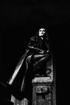 a woman sitting on top of a chest in a black dress and leather coat, with her hands behind her back