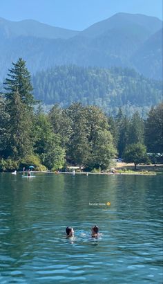 two people swimming in a lake with mountains in the background and trees on both sides