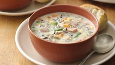 a bowl of soup on a white plate with a spoon and bread in the background