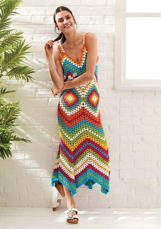 a woman standing in front of a white brick wall wearing a multicolored crocheted dress