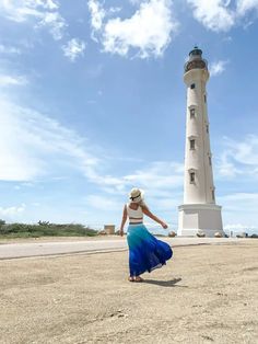 a woman in a blue dress and hat walking towards a light house on the beach