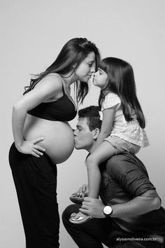 a pregnant woman kisses her husband's belly as they sit on a stool with their child