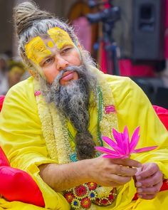 a man with a long beard and painted face sitting on a red chair holding a pink flower