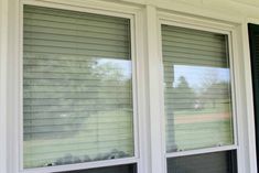 two windows that have blinds on them in front of a house with grass and trees behind them