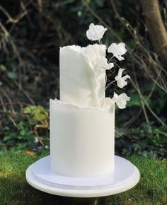 a white wedding cake with flowers on top sitting in the grass near some bushes and trees