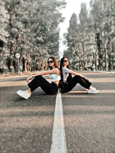 two young women sitting on the side of an empty road with trees in the background