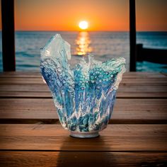 a glass vase sitting on top of a wooden table next to the ocean at sunset