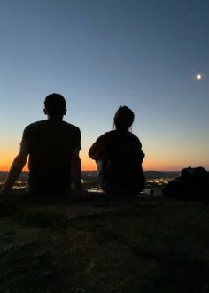 two people sitting on top of a hill at sunset