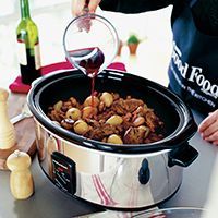 a person pouring red wine into a slow cooker filled with cooked meats and vegetables