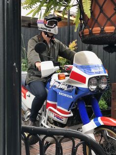 a man riding on the back of a red, white and blue dirt bike next to a planter