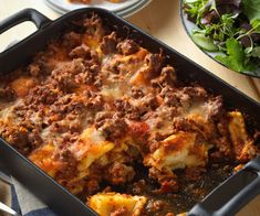 a casserole dish with meat and cheese in it sitting on a table next to a salad