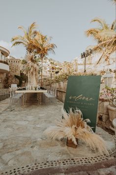 a sign sitting on top of a stone floor next to palm trees and chairs in front of a building