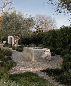 a stone bench sitting in the middle of a gravel area surrounded by bushes and trees