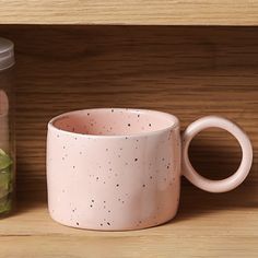 a pink coffee cup sitting on top of a wooden shelf next to a jar filled with cucumbers