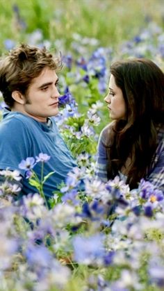 a man and woman sitting in a field of flowers looking into each other's eyes