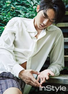 a young man sitting on a wooden bench in front of some bushes and trees looking down at his watch