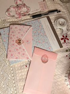 some pink and blue envelopes are laying on a table with lace, flowers and other items