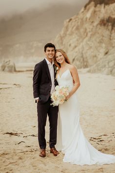 a bride and groom pose for a photo on the beach in front of some cliffs