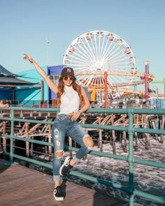 a woman is posing on a pier with her arms in the air while wearing ripped jeans and a baseball cap