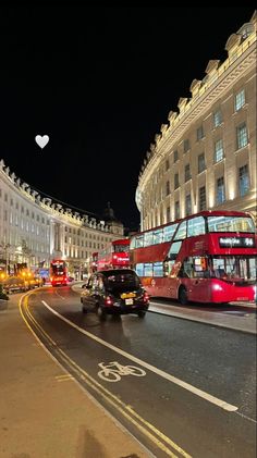 two red double decker buses driving down a street next to tall white buildings at night