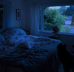 a bed in a dark room with blue light coming through the window and trees outside