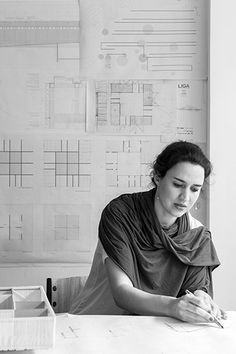 a woman sitting at a table writing in front of a wall with plans on it