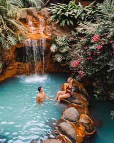 two people in a pool with waterfall and water fall behind them, surrounded by greenery