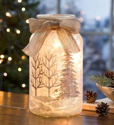 a mason jar filled with snow and pine cones on top of a wooden table next to a christmas tree