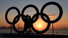 two people standing next to each other in front of the olympic rings at sunset with mountains in the background