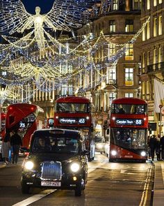 two red double decker buses driving down a street with christmas lights on the buildings behind them