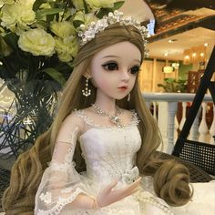 a doll wearing a white dress and tiara sitting on a table with flowers in the background