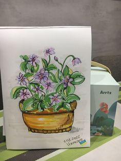 a painting of purple flowers in a yellow pot on a table next to a card