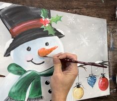 someone is painting a snowman with christmas ornaments on the board and holding a paintbrush