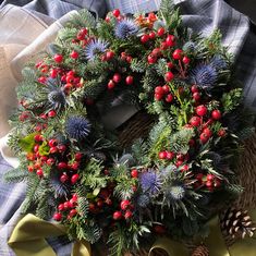 a christmas wreath with berries and pine cones on a plaid table cloth, next to evergreens