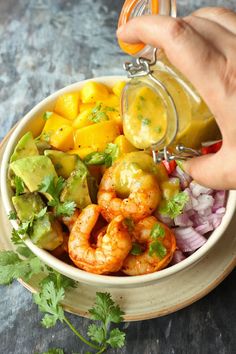 a person pouring dressing into a bowl filled with shrimp, avocado and mango salsa